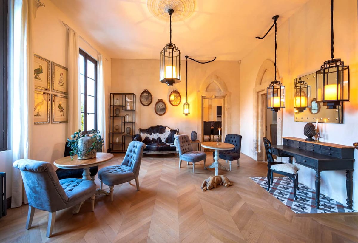 Wide interior shot of a furnished salon or reception room within what appears to be a French château or historic manor. The space features herringbone parquet wood flooring, high ceilings with an ornate plaster ceiling medallion, and arched Gothic-style doorways. Lighting comes from multiple black iron cage-style pendant lanterns with warm amber glow, plus a candle on a dark console table along the right wall. Seating includes tufted velvet armchairs in grey-blue tones, a cowhide canapé sofa, and several small round wooden pedestal tables. Decorative elements include framed botanical bird prints arranged in a grid on the left wall, oval-framed portrait photographs on the back wall, a dark metal open shelving unit displaying ceramic objects, and a patterned geometric tile rug in grey, white, and red. A golden retriever dog rests on the floor in the center of the room. The overall styling mixes antique French furniture with industrial lighting details, creating an eclectic, character-rich interior. Potential venue feature image.