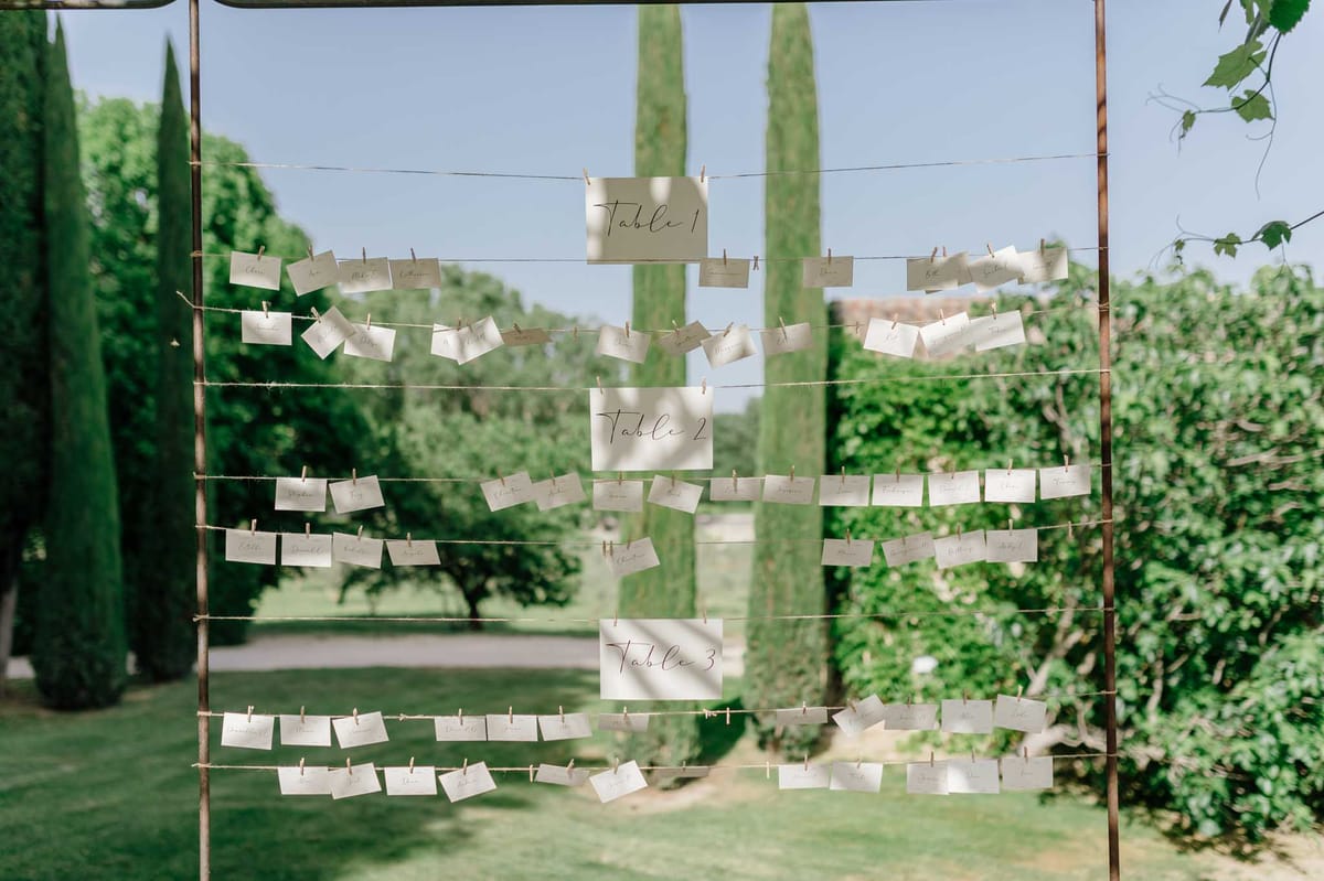 Outdoor seating chart display with cream cards clipped to wire strands between wooden poles, cypress trees behind