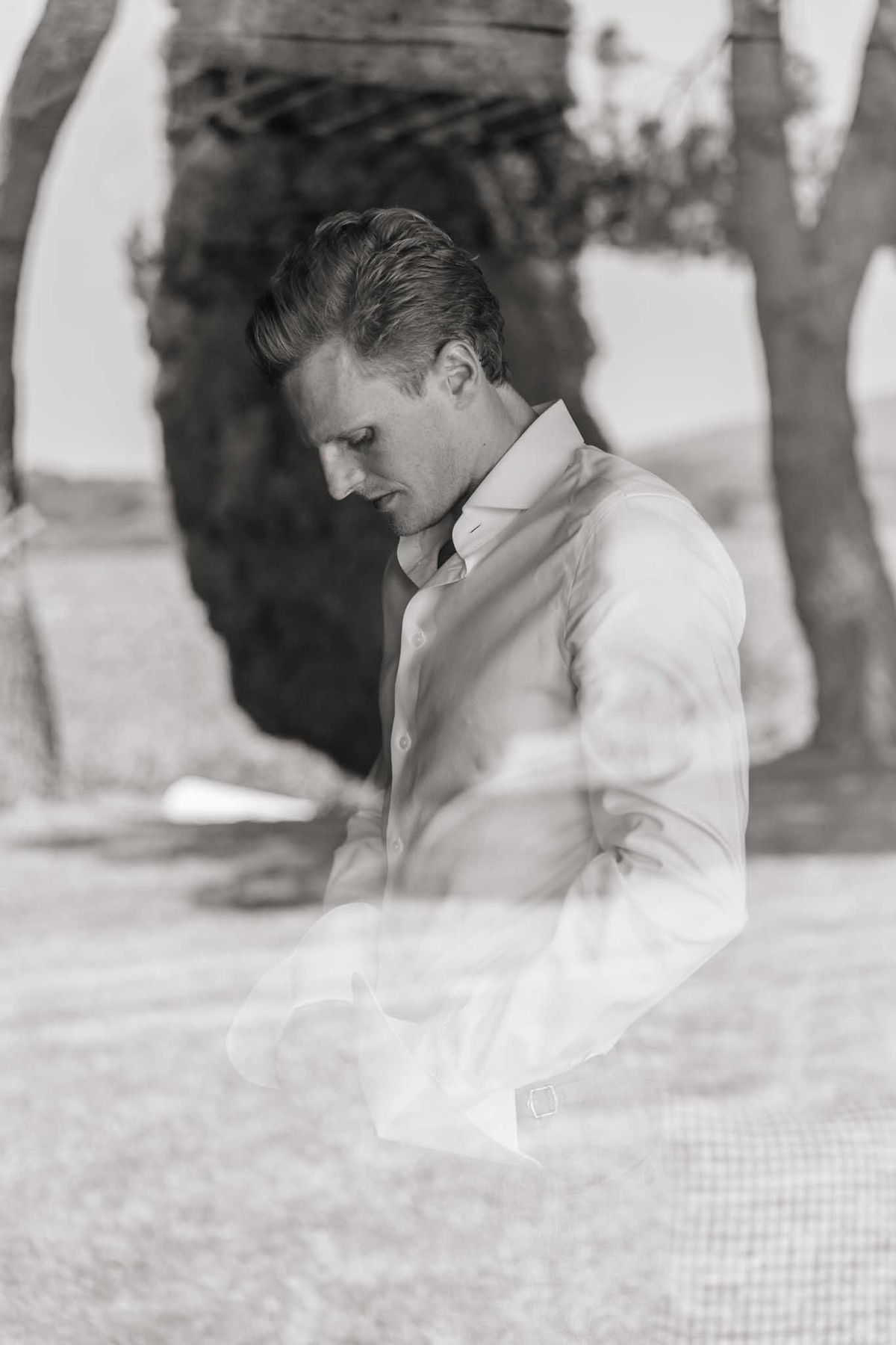 Groom in white dress shirt looking down in quiet contemplation during the ceremony, stone archway visible behind.