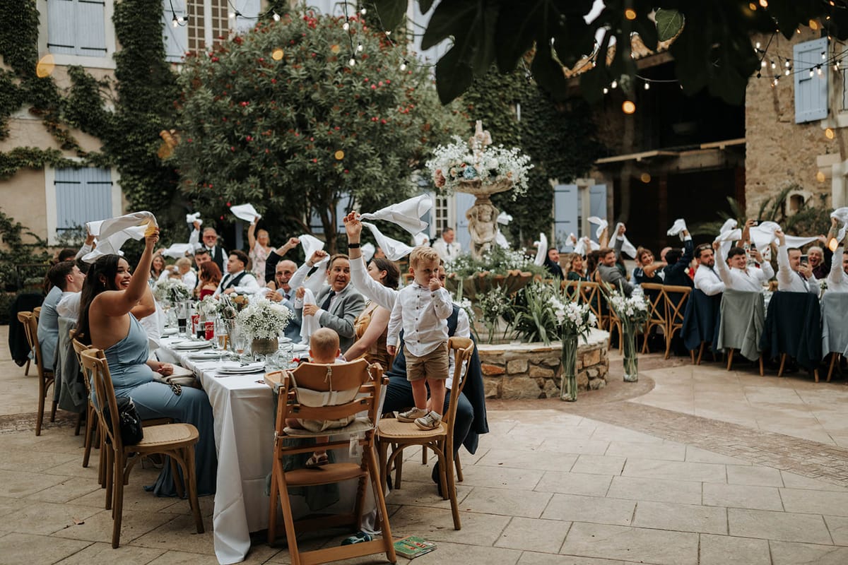 Wedding guests wave white napkins at long banquet tables in ivy-covered courtyard with string lights overhead