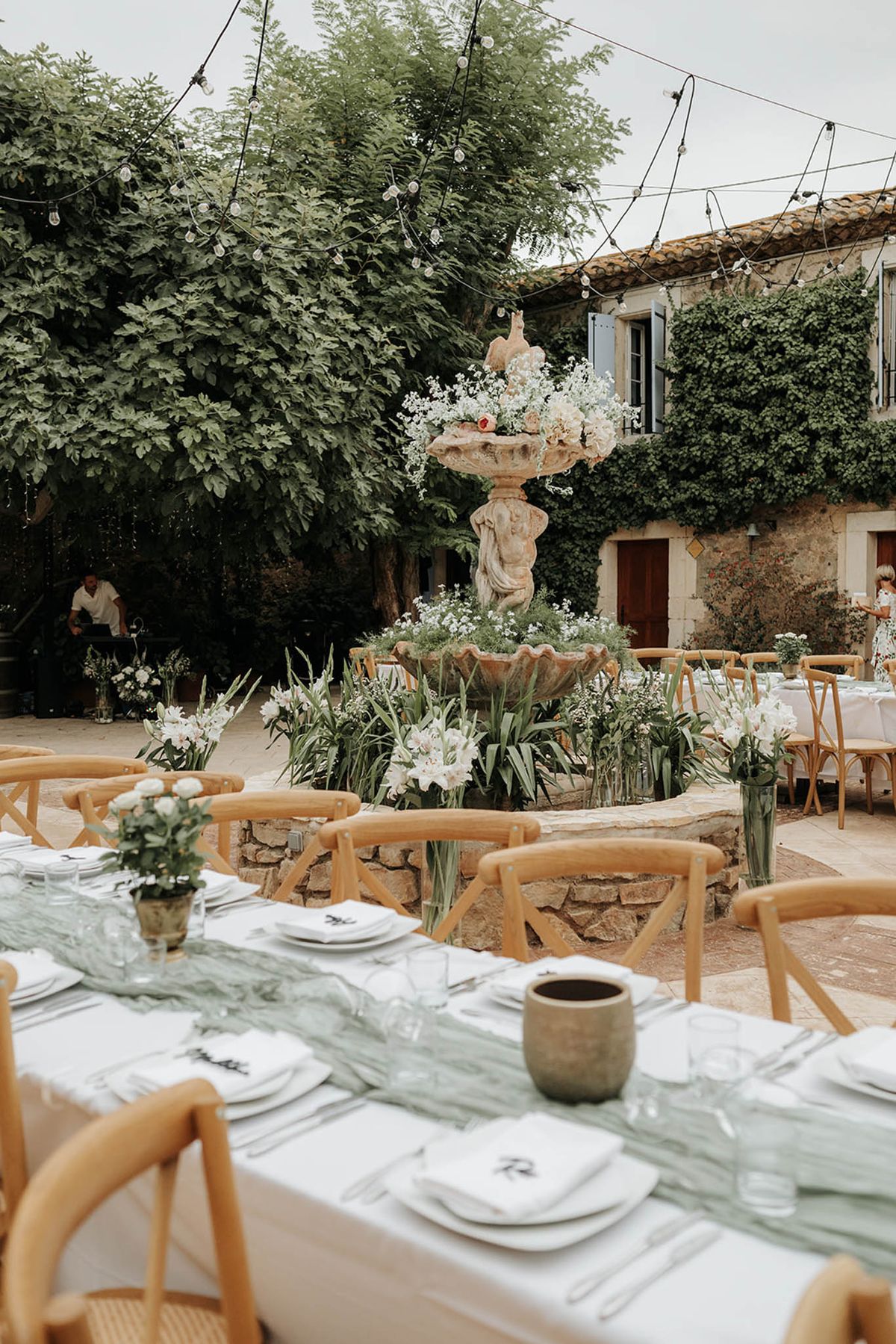 Provencal courtyard reception with stone fountain, sage runners, white roses, and Edison string lights