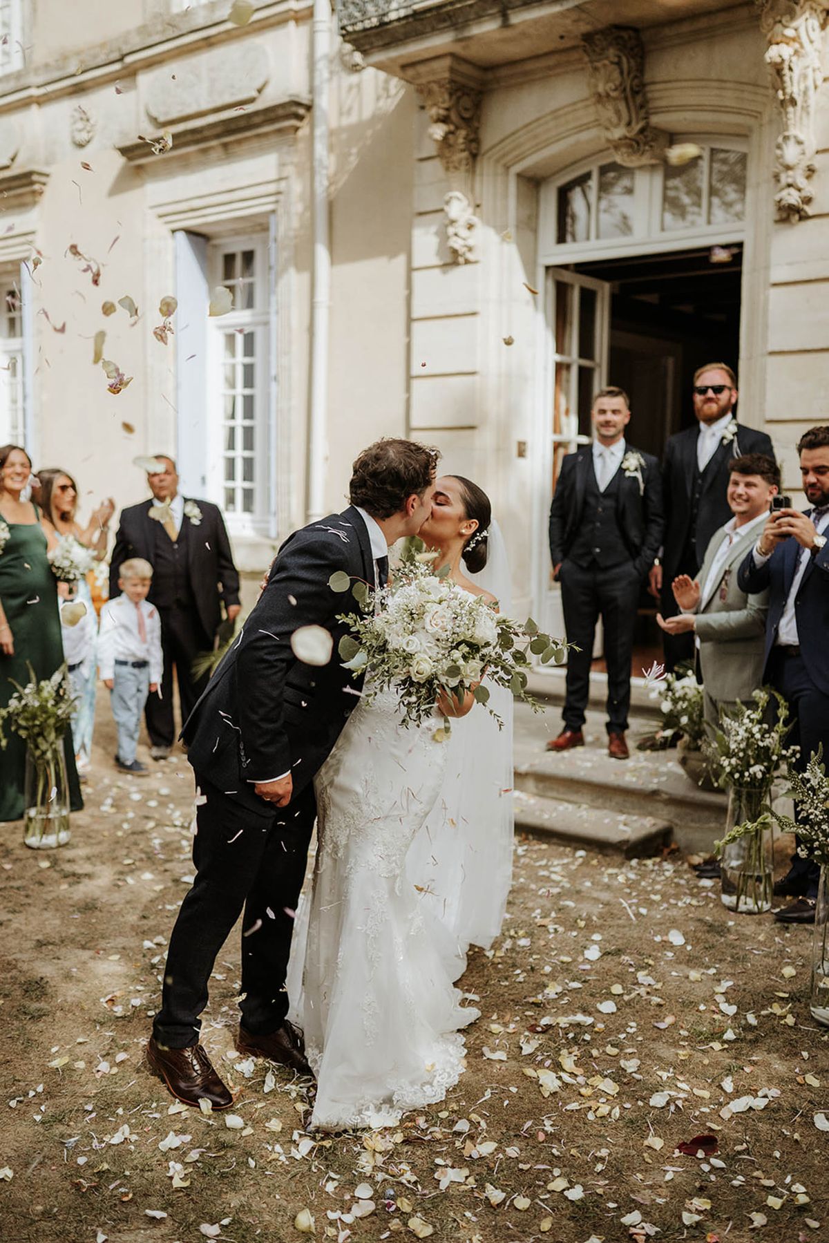 Bride in ivory lace gown and groom kissing as guests throw dried petal confetti before chateau facade