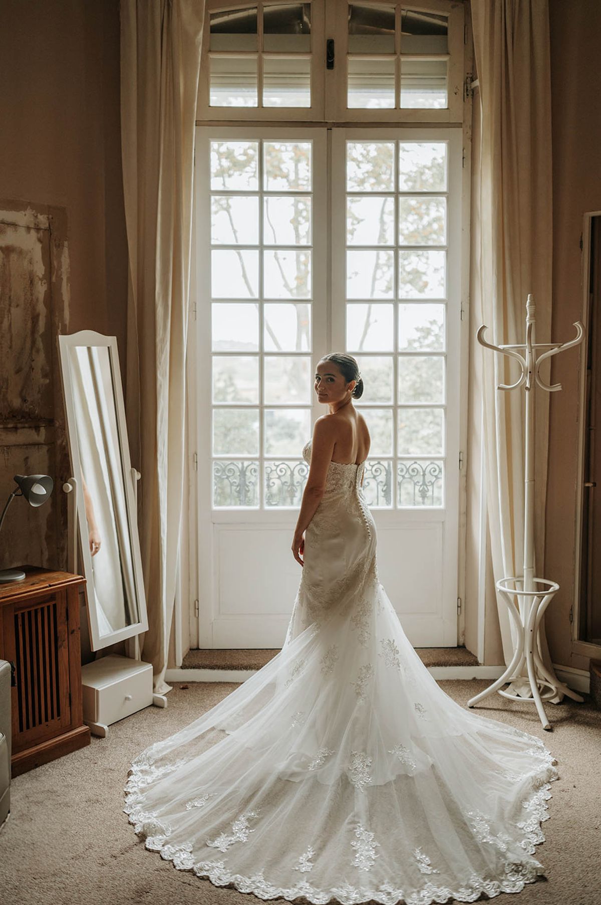 Bride in strapless lace mermaid gown with cathedral train looking over her shoulder in front of tall French doors
