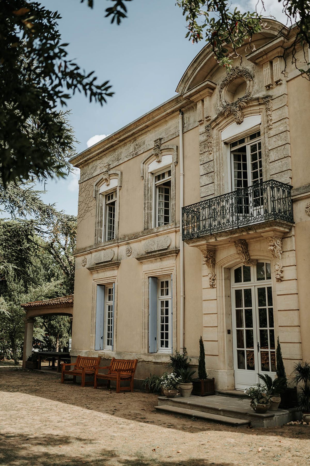 Exterior shot of a two-storey French château with cream-rendered stone façade featuring ornate baroque-style carved stonework, a wrought-iron juliet balcony on the upper floor, and pale blue shutters on the ground-floor windows. The main entrance has white French doors flanked by carved stone pilasters and accessed via a few stone steps, with potted cypress trees and flowering plants placed at the entrance. Two teak garden benches are positioned along the front of the building, and a covered pergola structure with a terracotta-tiled roof is visible to the left. The foreground shows a gravel courtyard, and the shot is framed by overhanging tree branches. Wide architectural shot with no people visible. Potential venue feature image.