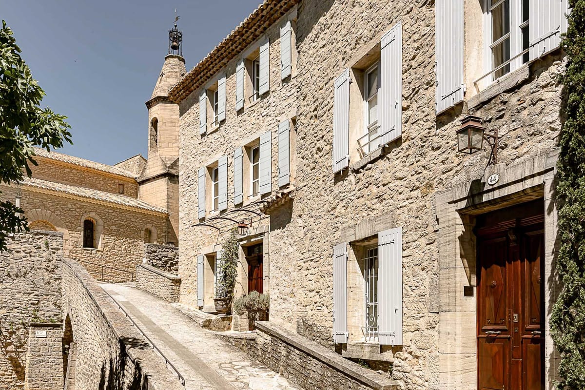 A wide-angle exterior shot of a traditional Provençal stone building in a French village, featuring warm golden limestone facades, pale grey-blue shutters, and solid dark wood entrance doors — including a double door numbered 44 with a wrought iron wall lantern. A sloped village lane runs alongside the building, bordered by a low stone wall with iron railing. In the background, a church bell tower with a weathervane is visible, along with the tiled rooftop of an adjacent stone chapel or church. Potted plants are placed at the ground-floor entrance. No people are present in the image. Potential venue feature image.