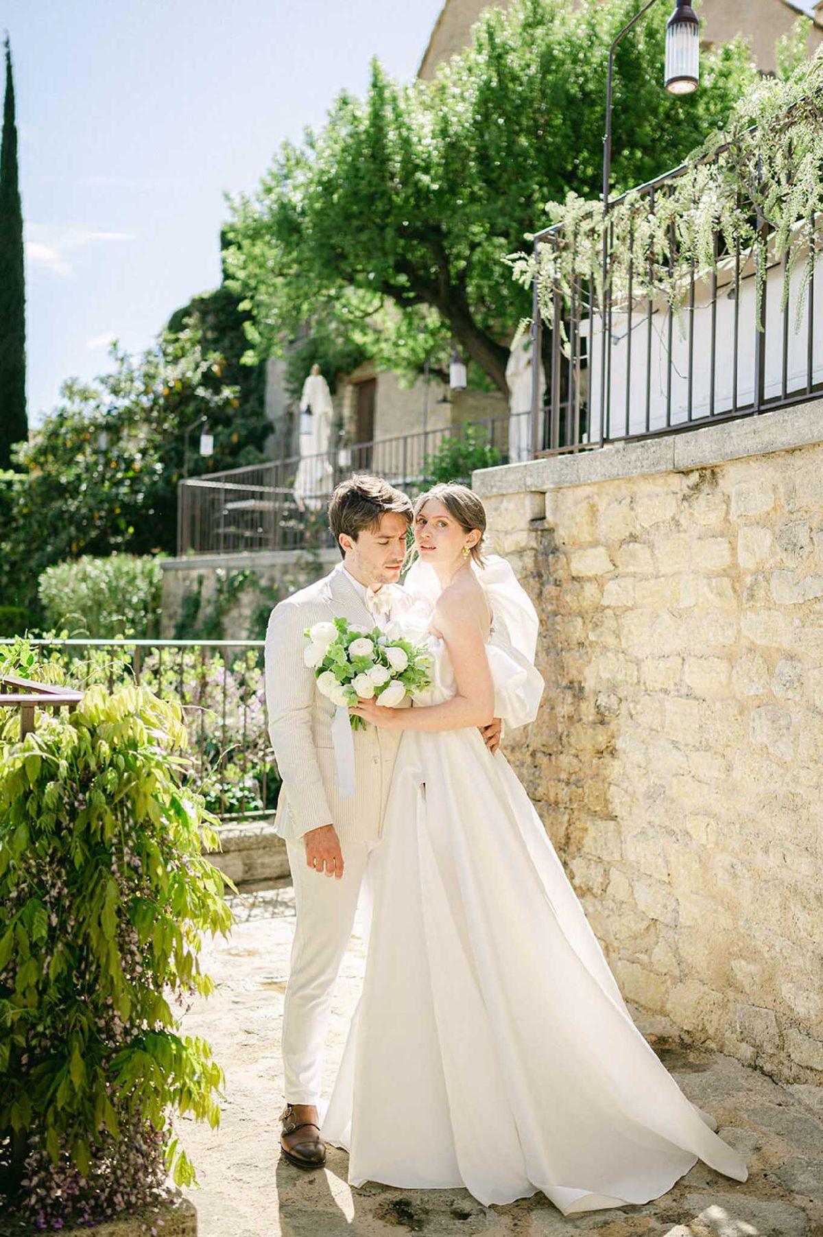 A couple portrait taken outdoors in the grounds of what appears to be a Provençal stone property, with iron railings, a terraced garden, and pale limestone walls visible in the background. The groom wears a light cream pinstripe suit with white trousers and brown leather loafers, while the bride wears a white ball gown with voluminous puff sleeves and a full skirt with a train. The bride holds a compact round bouquet of white garden roses and white ranunculus with green foliage. The groom stands behind the bride with his arm around her waist, his head tilted toward hers while she looks back toward the camera. The shot is a medium full-length portrait taken in bright midday sunlight.