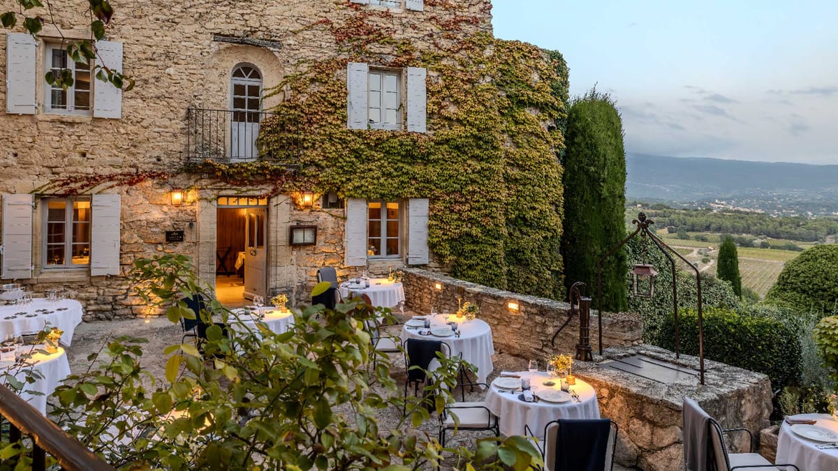 An outdoor terrace dining setup at dusk, arranged against the façade of a large ivy-covered stone building with white shutters and warm interior lighting visible through open doors and windows. Approximately six to eight round tables are dressed in white floor-length linens, set with glassware, plates, and small candlelit centerpieces with what appear to be small yellow floral arrangements. Black metal chairs accompany each table. Wall-mounted lanterns and ground-level lights illuminate the terrace, creating warm ambient lighting in the early evening. A wrought-iron staircase railing with a lantern post is visible to the right, and the terrace overlooks a wide valley with vineyard rows visible in the distance. Wide-angle shot emphasizing the full setting. Potential venue feature image.