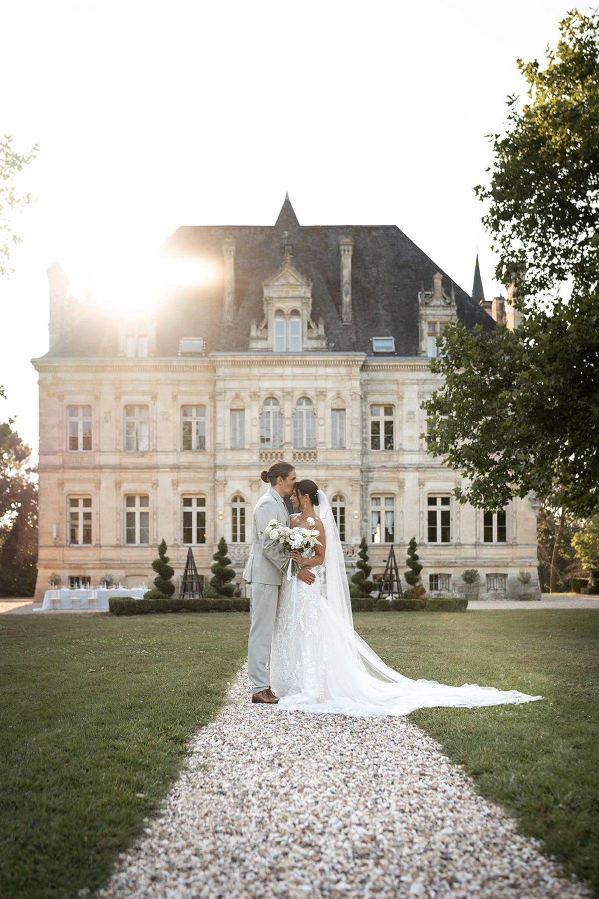 Bride and groom sharing a forehead kiss on a gravel path in front of a classical French chateau at golden hour
