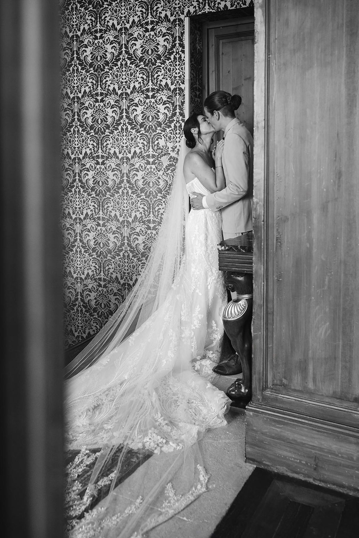 Black and white photo of bride and groom kissing in narrow hallway with baroque wallpaper