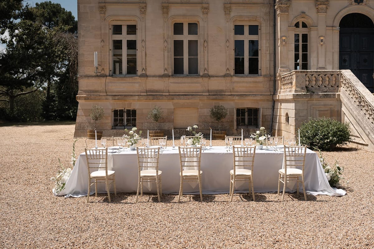 Single long banquet table on gravel courtyard in front of honey-coloured château with white hydrangea centerpieces