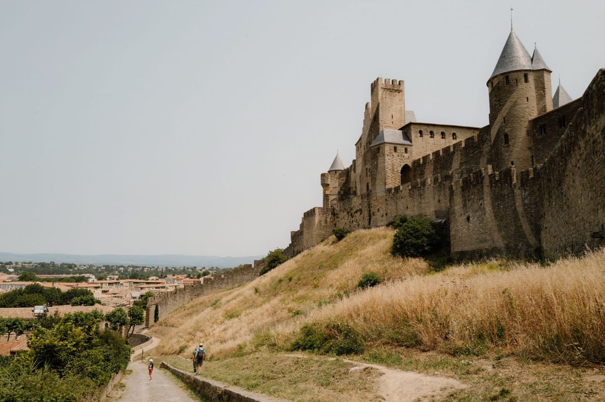 A wide-angle exterior shot of the medieval fortified city of Carcassonne in southern France, showing the iconic stone ramparts, battlements, and conical-roofed towers rising above a dry grassy embankment. Two visitors — one wearing a blue cap and backpack, another in red — walk along a path that runs alongside the outer walls, with the lower town's terracotta-roofed buildings visible in the background. This image does not appear to contain any wedding-related subjects, decor, or activity. Potential venue feature image.
