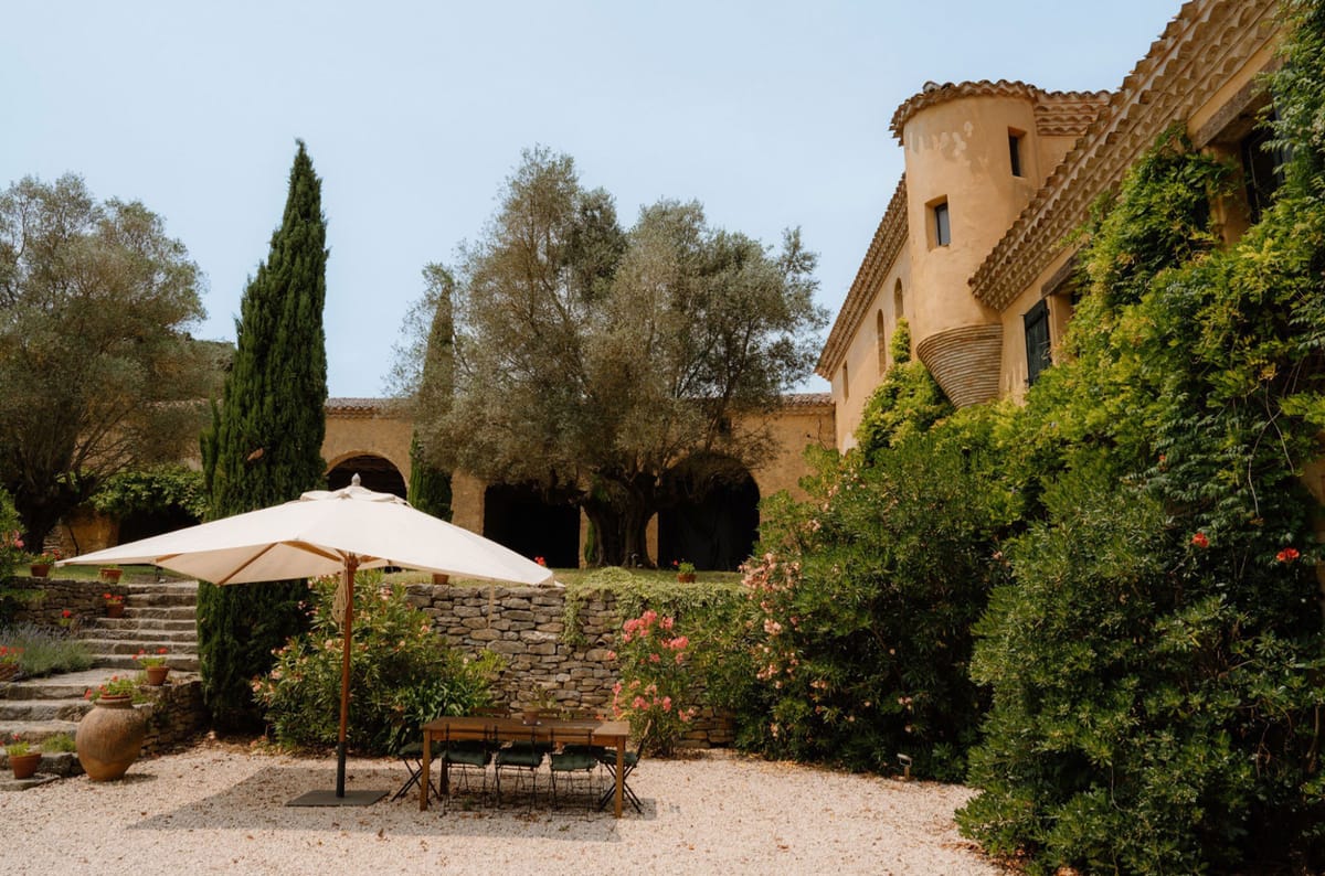 An outdoor wide shot of a Provençal-style mas or bastide venue featuring a warm ochre-toned stone building with a cylindrical tower, terracotta roof tiles, and arched colonnades visible in the background. The grounds include a gravel courtyard with a cream market umbrella shading a small wooden table and dark metal folding chairs, set up as a seating area. Dry-stone retaining walls, terracotta pots with red geraniums, pink oleander bushes, cypress trees, and climbing foliage cover the building facade, contributing to the overall rustic Provençal aesthetic. No people are present in this image. Potential venue feature image.