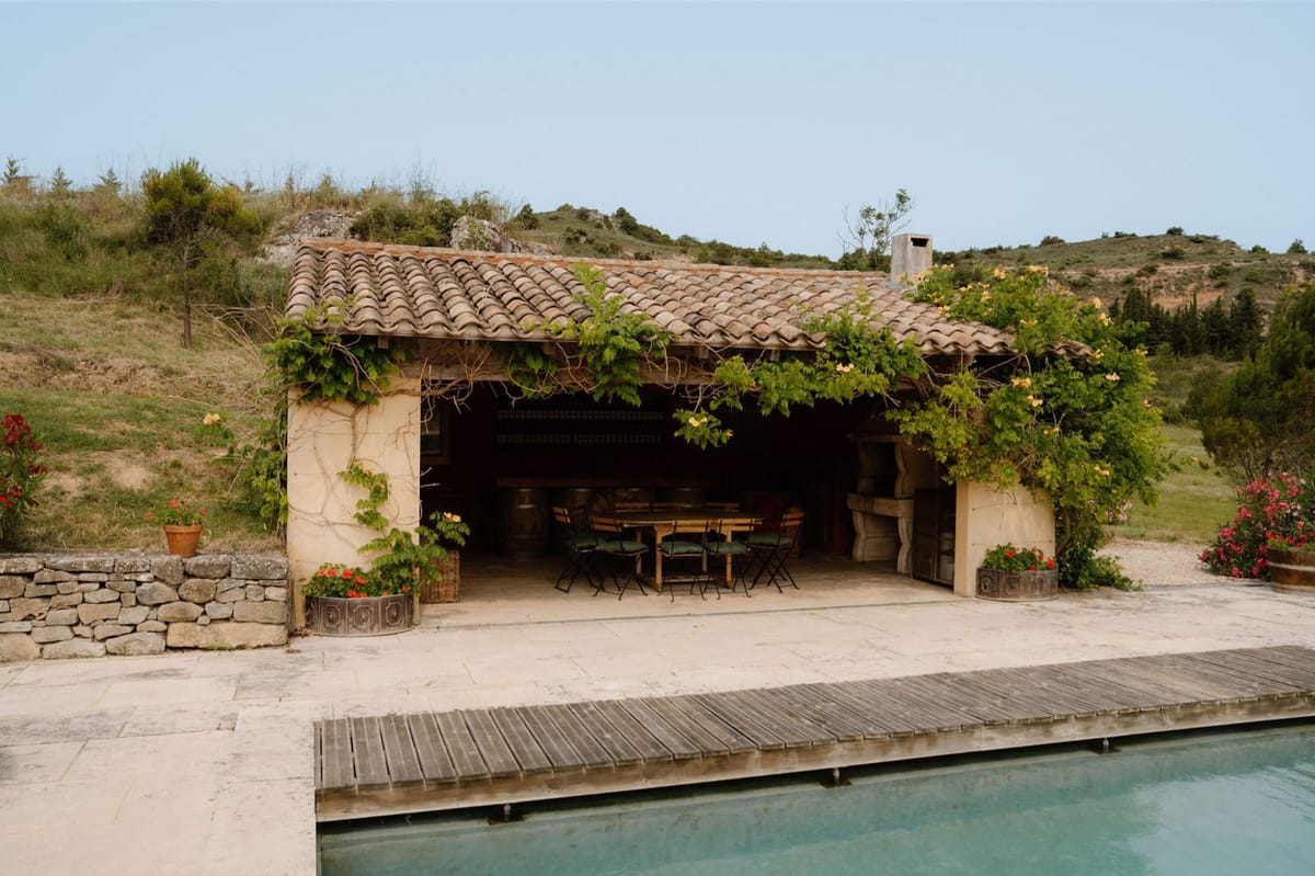 A wide shot of an outdoor pool house or covered dining pavilion at what appears to be a rural French property. The structure features a traditional terracotta tile roof with climbing vines and yellow flowering plants across the facade, and open-fronted walls revealing a rustic interior with a long wooden dining table surrounded by folding chairs with dark green cushions. Large decorative barrel-style planters flank the entrance, filled with red flowering plants. A stone-paved terrace connects the pavilion to a rectangular swimming pool in the foreground, with a wooden slatted bridge or cover across part of the water. The setting has a rustic Provençal character with dry-stone retaining walls and hillside terrain in the background. No people are present in the image. Potential venue feature image.