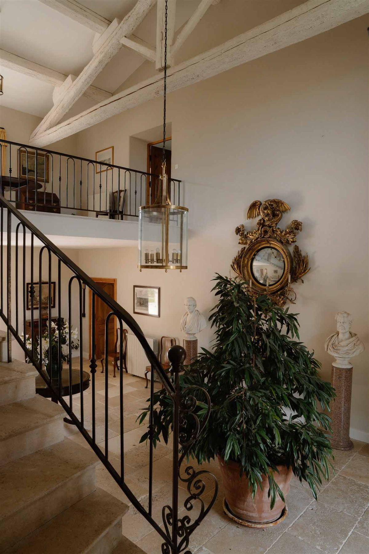 A wide interior shot of a French country house entrance hall, showing no people or wedding activity. The space features a curved wrought-iron staircase with ornate scrollwork, whitewashed exposed ceiling beams, and large stone tile flooring. A brass and glass lantern-style pendant light hangs from the ceiling. Decorative elements include a gilded convex mirror with an elaborate eagle and rococo-style carved surround, two white marble classical busts on stone pedestals, a large green potted plant in a terracotta urn, and framed artwork on the walls. A mezzanine level with a black iron railing is visible above. The overall interior style is classic French provincial with antique furnishings. Potential venue feature image.