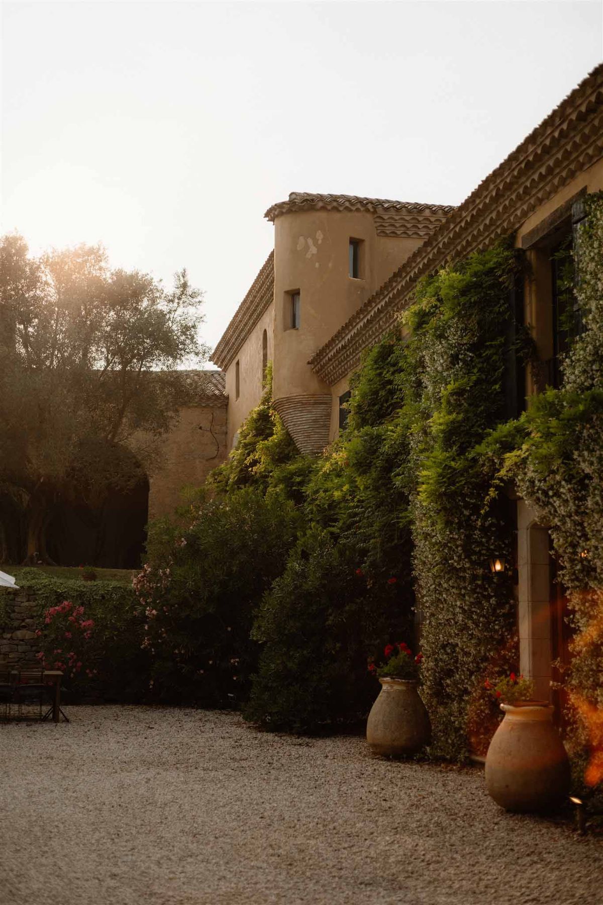 Ochre Provencal bastide with rounded tower, climbing vines, and terracotta urns on gravel at dusk