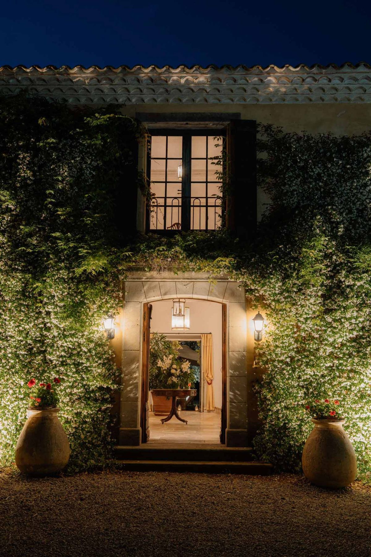 A nighttime exterior shot of a historic mas or domaine-style venue, showing the main entrance facade lit by warm wall-mounted lanterns and a hanging pendant light framing the open arched doorway. The two-story stone building is heavily covered in climbing vines with small white blooms, illuminated by uplighting that highlights the texture of the greenery against the pale render. Two large terracotta urns planted with red geraniums flank the entrance on a gravel forecourt. Through the open double doors, a warmly lit interior is visible with a dark wood console table, cream curtains, and white floral arrangements. A tall iron-framed window on the upper floor is backlit from within, revealing a pendant light inside. The wide architectural shot is taken straight-on, centered on the doorway. Potential venue feature image.
