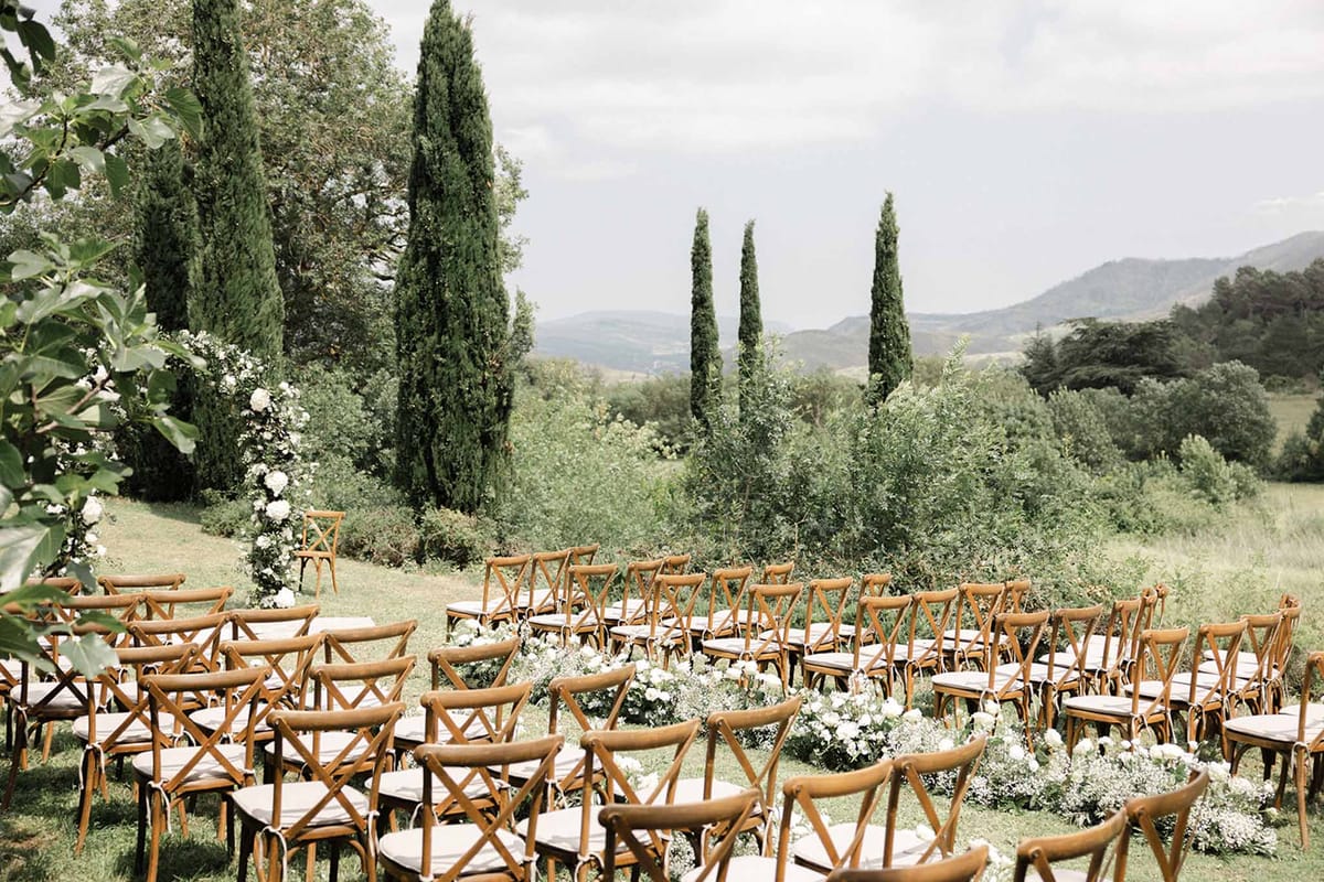 Outdoor ceremony setup with crossback chairs and cream cushions on lawn, white floral aisle markers and cypress tree backdrop