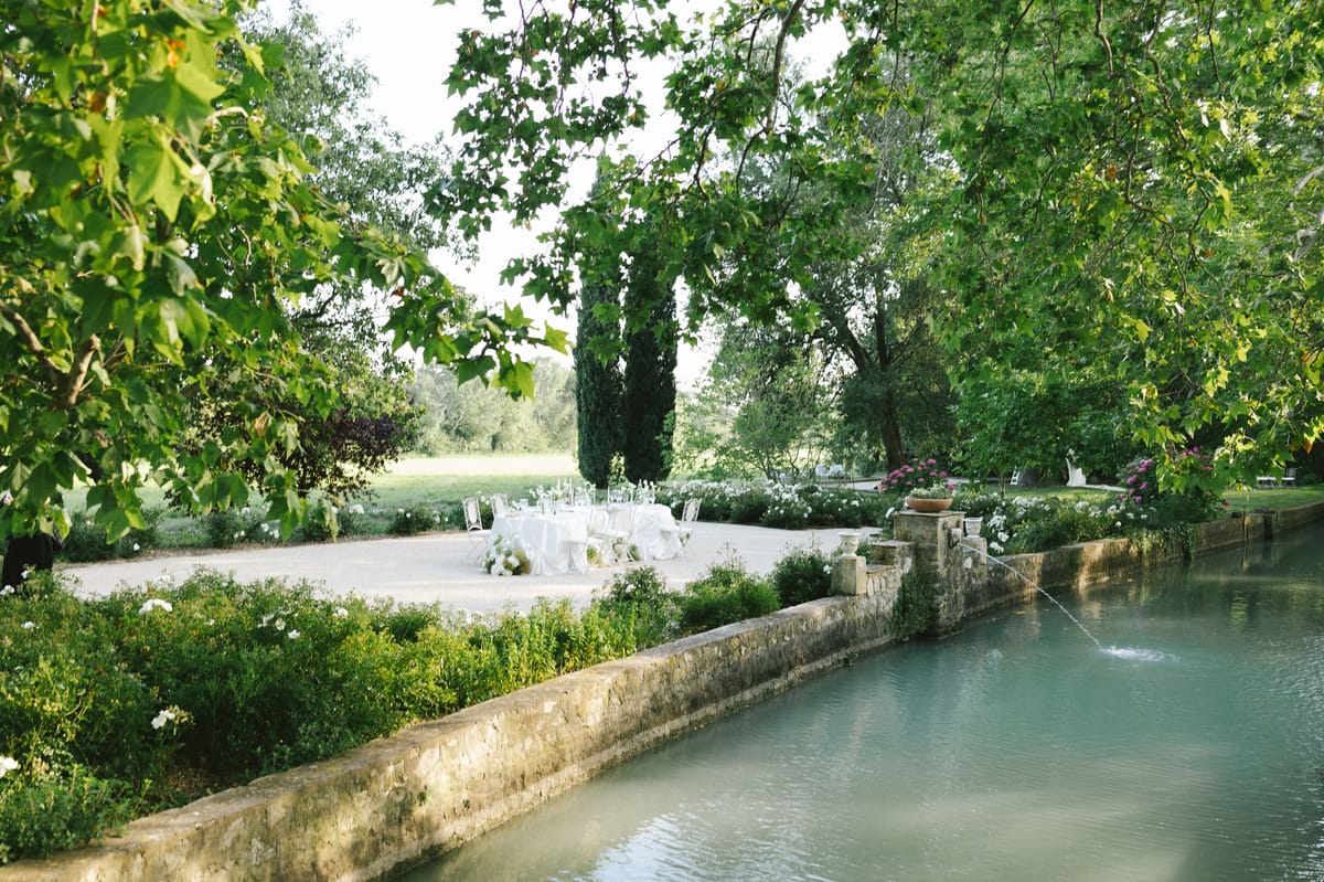 Outdoor reception table set for dinner beside ornamental canal with fountain, white roses, and cypress trees in garden