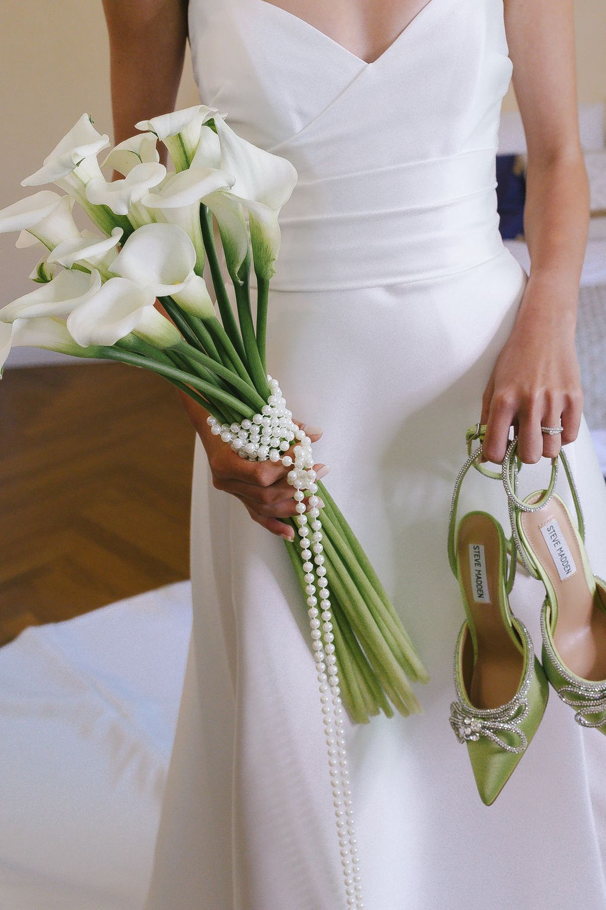 Close-up of bride's hands showing white calla lily bouquet with pearl wrap and sage green crystal-embellished heels