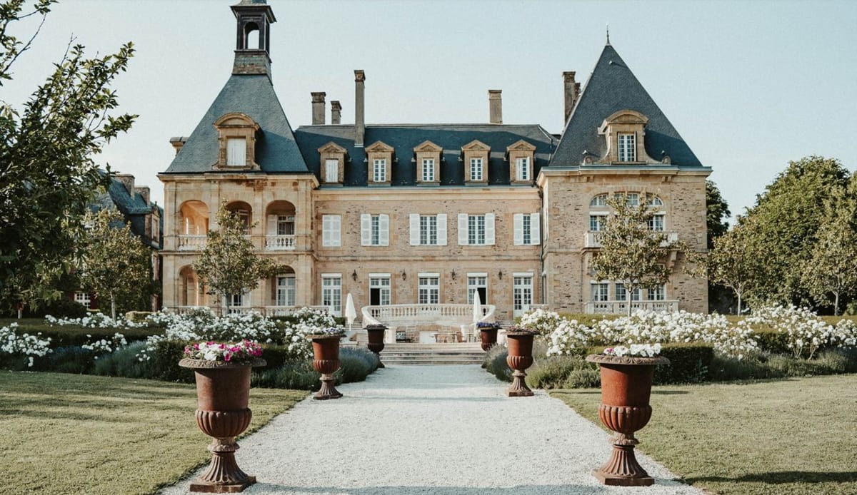 Wide-angle exterior shot of a large French château with warm honey-toned stone facades, slate-grey mansard roofs with pointed corner turrets, and multiple dormer windows with white shutters. A symmetrical gravel driveway leads from the foreground to a balustraded stone terrace at the entrance, flanked by four large terracotta urn planters filled with pink and white flowering plants. White rose borders line both sides of the approach path, and closed white parasols are visible on the terrace alongside seating. No people are present in the image. Potential venue feature image.