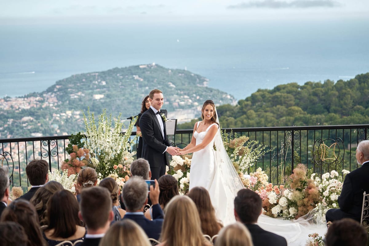 An outdoor ceremony taking place on an elevated terrace with a sweeping panoramic view of the French Riviera coastline and a hillside town in the background. The bride wears a fitted ivory gown with a structured square neckline and a long flowing veil, while the groom is dressed in a black tuxedo with a black bow tie and a white boutonniere; the couple holds hands facing their guests. A female officiant stands behind them holding a ceremony booklet, flanked by lush floral arrangements featuring white delphinium, ivory and blush roses, peach spray roses, and dried pampas-style grasses forming a ground-level floral display along the terrace railing. A gold monogram decorative element is visible on the wrought-iron railing to the right. Approximately 20–30 seated guests are visible in the foreground, and the wide-angle shot captures both the ceremony in full and the dramatic coastal backdrop. Potential venue feature image.