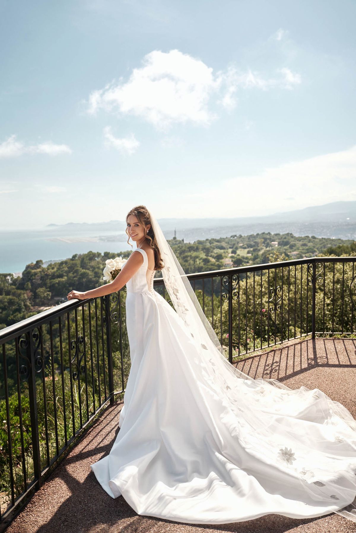 A bridal portrait taken outdoors on a terrace with wrought-iron scrollwork railings, overlooking a panoramic coastal landscape with a bay visible in the distance. The bride stands in three-quarter profile, glancing back toward the camera, wearing a white sleeveless A-line gown with a low back and an extended cathedral-length train that spreads across the terrace floor. She wears a long cathedral veil with scattered appliquéd floral details throughout its length. Her bouquet consists of white roses and she wears her dark hair in a half-up style. The overall styling is classic and clean, with a modern minimalist dress paired with an ornate veil. Full-length portrait shot in bright natural daylight.