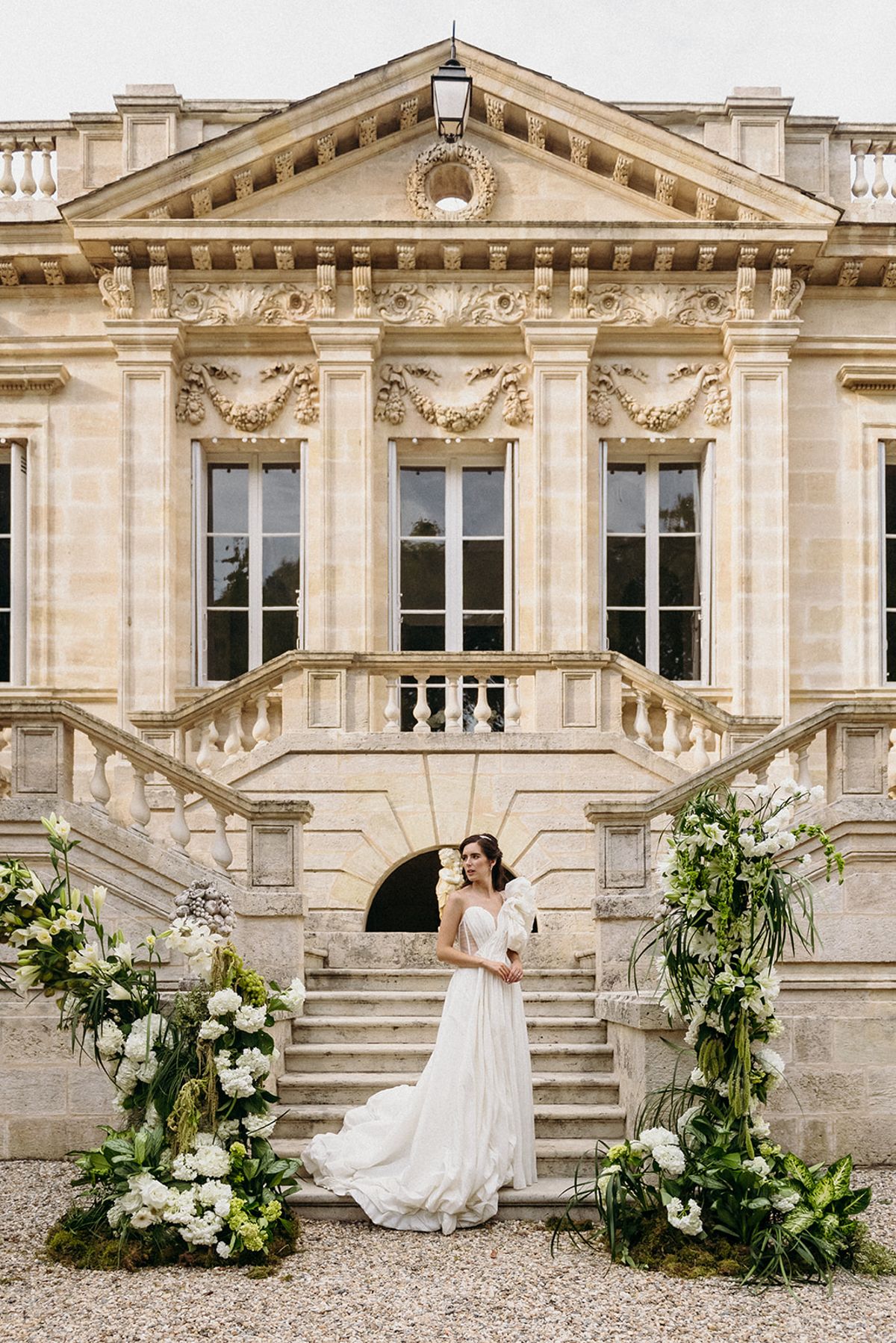 A bridal portrait taken outdoors in front of a French château featuring ornate classical stone architecture with a pediment, carved decorative friezes, tall arched windows, and a double-sided stone staircase with balustrade. The bride stands alone at the base of the staircase on a gravel forecourt, wearing a white A-line gown with a sweetheart neckline, a sheer tulle skirt with a trailing train, and a large off-shoulder bow detail. Large floral arrangements frame her on both sides, composed of white hydrangeas, white lilies, white stock flowers, and lush dark green foliage including tropical leaves and tall grasses, creating a structured yet organic ceremony backdrop. The composition is a wide, symmetrical portrait shot with the château façade filling the background. Potential venue feature image.
