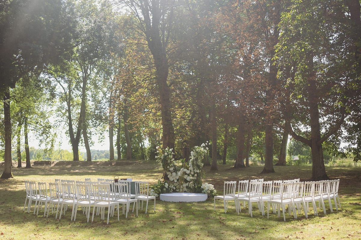 An outdoor ceremony setup photographed in wide shot before guests arrive, set on a lawn within a wooded parkland area. White Napoléon chairs are arranged in a semicircular layout on either side of a central aisle, facing a circular white platform used as an altar. The altar is decorated with a large, lush arrangement of white florals — appearing to include roses, peonies, and greenery — clustered at the base and framing the ceremony space. The overall decor palette is white and green, with a classic, garden-party aesthetic. Soft backlighting filters through the tree canopy, casting dappled light across the setup. Potential venue feature image.