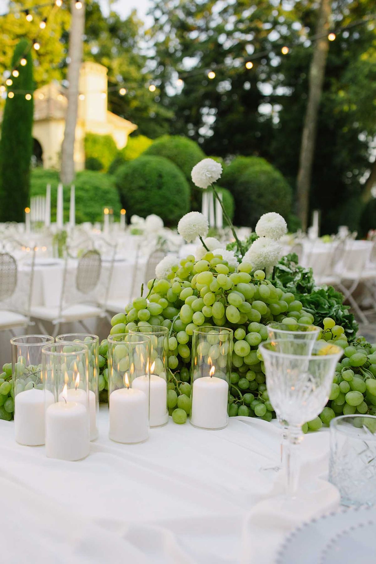 Centerpiece with cascading green grapes, white hydrangeas and pillar candles, cypress trees and stone building behind