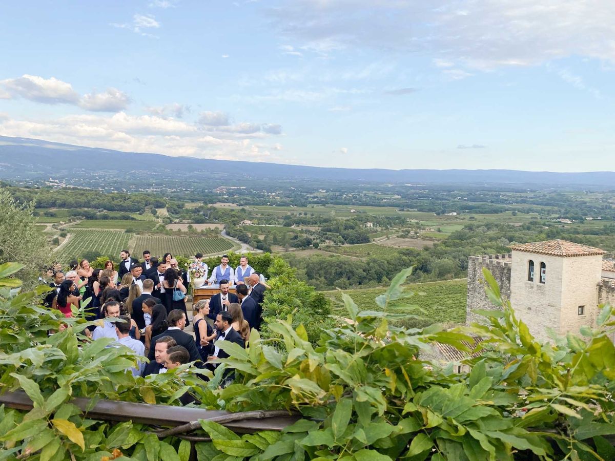An outdoor cocktail hour taking place on a high terrace or rampart of a stone château, with approximately 30–40 guests mingling in the late afternoon light. Most guests are dressed in black tie — men in dark suits or tuxedos with bow ties, women in black cocktail dresses — with a few in lighter colors including one guest in a red dress. A bar or serving table with a floral arrangement featuring peach and pink tones in a large urn is visible at the center of the group. To the right, a medieval stone tower with a crenellated parapet and terracotta-tiled roof is visible as part of the château structure. The wide shot is taken from behind a foreground of green climbing foliage, looking down slightly onto the gathering, with an expansive valley of vineyards and agricultural fields stretching to a distant mountain range in the background. Potential venue feature image.