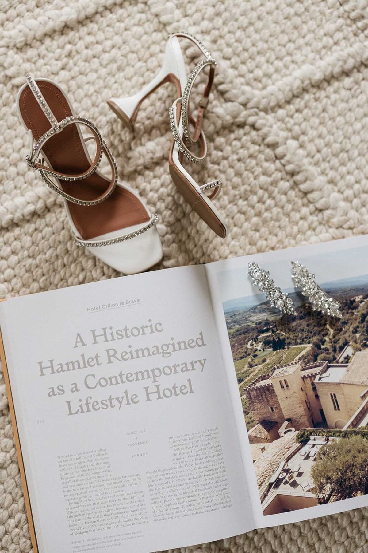 Flat lay of crystal-strap bridal sandals, drop earrings, and open venue book on cream knit textile
