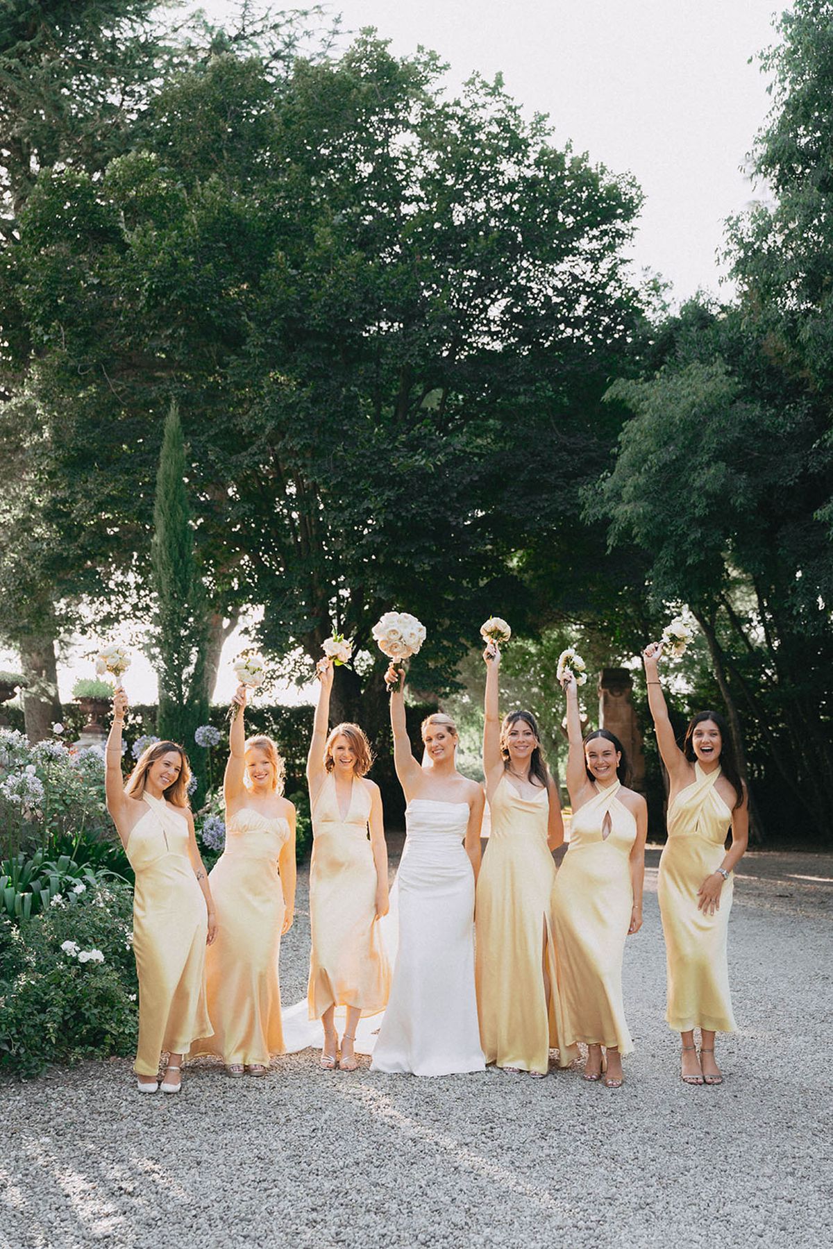 Bride in ivory column gown with seven bridesmaids in pale yellow dresses raising bouquets in tree-lined garden