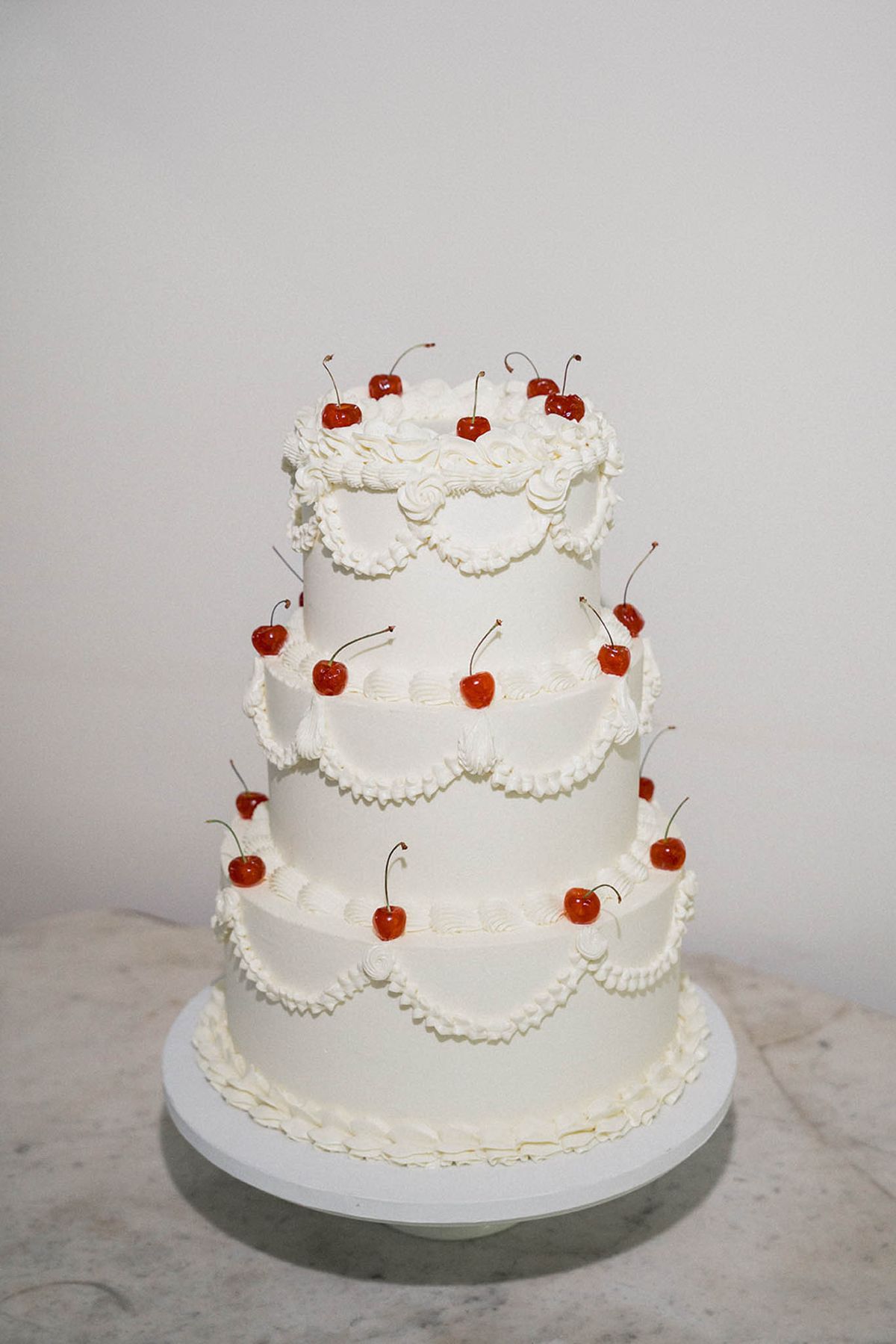 Four-tiered ivory buttercream wedding cake with scalloped piping and red maraschino cherry decorations on marble surface