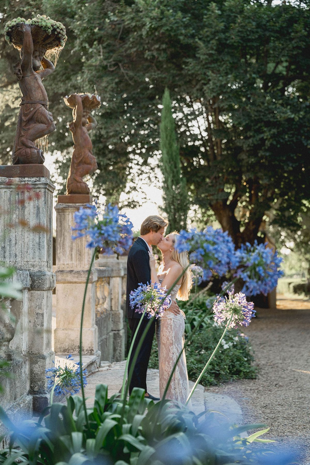 Bride and groom kissing on garden pathway lined with blue delphiniums and stone urns