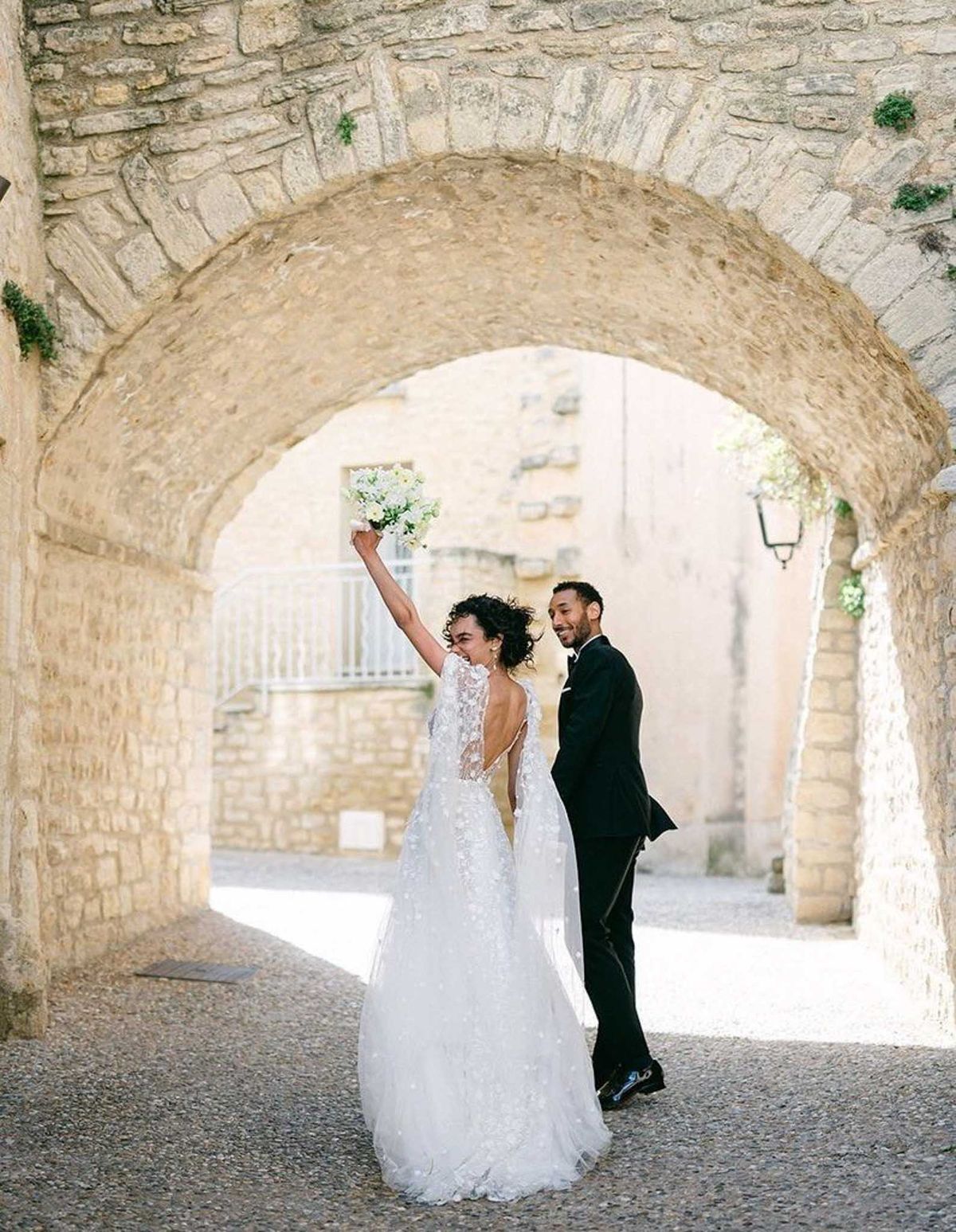 Bride in lace applique gown and groom in tuxedo celebrate under stone archway in French village