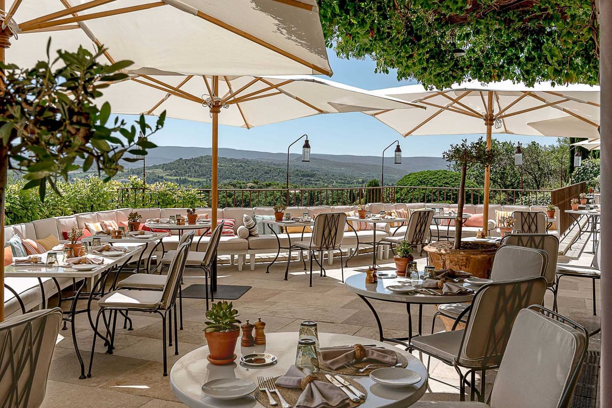 Provencal terrace with bistro tables, white parasols, and striped banquette cushions overlooking hillside