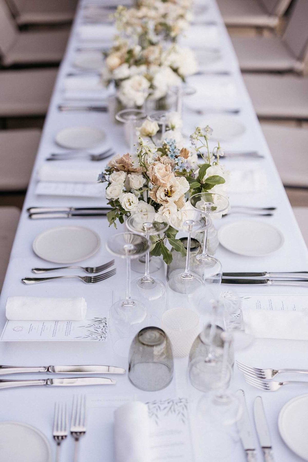 A close-up, slightly angled shot of a long reception dining table set for a wedding, with no people present. The table is covered in a pale ice-blue linen and laid with matte white round plates, white folded napkins, silver flatware, and clear stemmed wine and coupe glasses. A series of low floral centerpieces run down the center of the table, featuring ivory roses, blush lisianthus, soft taupe garden roses, pale blue forget-me-nots, and loose green foliage arranged in small clear glass vessels. Printed menus with a delicate botanical illustration are placed at each setting. The overall palette is white, ice blue, blush, and soft taupe, creating a refined, minimal modern aesthetic. The composition is a perspective detail shot looking down the full length of the table.