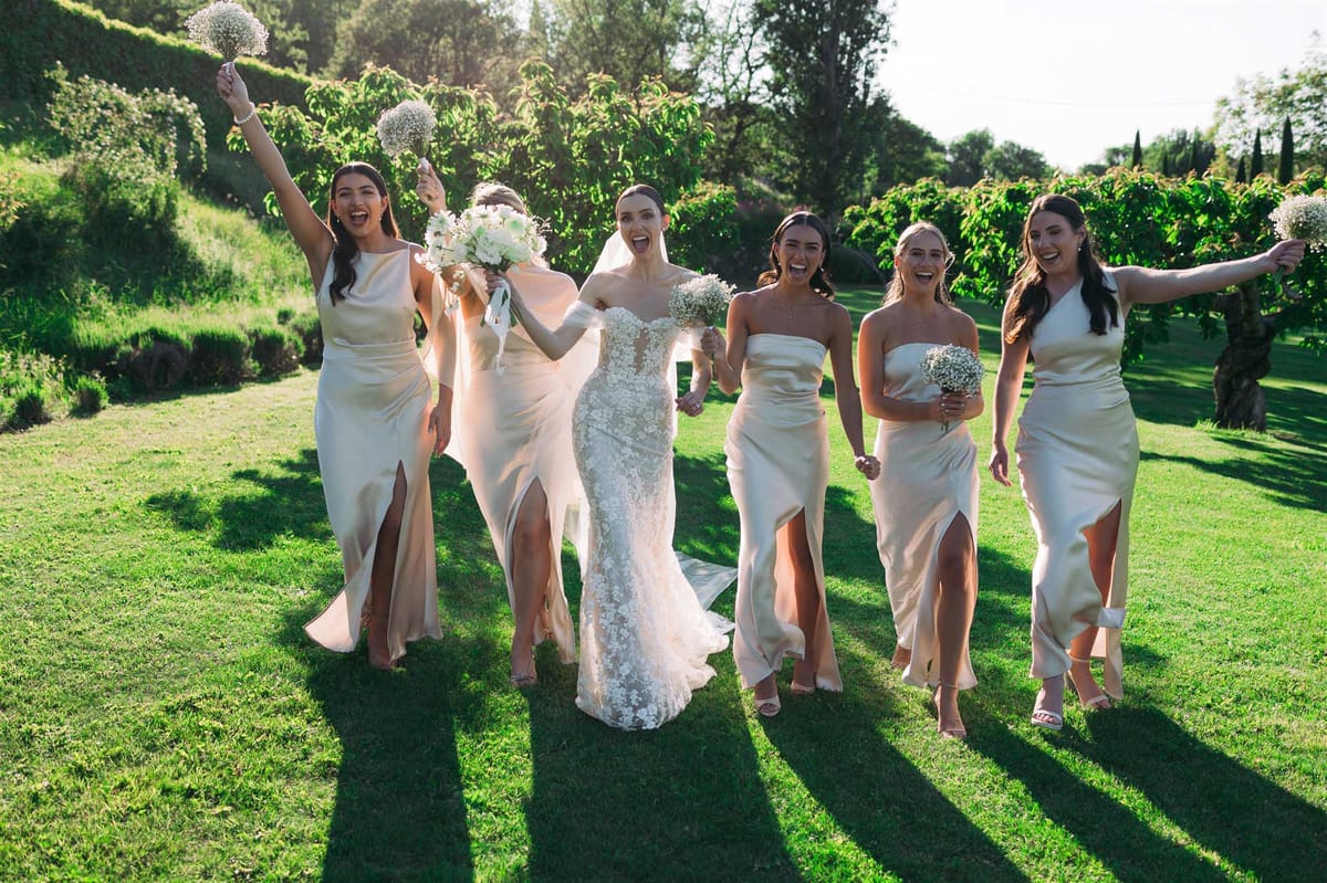Bride in beaded ivory gown with five bridesmaids in cream champagne slip dresses on lawn with cypress hedgerows