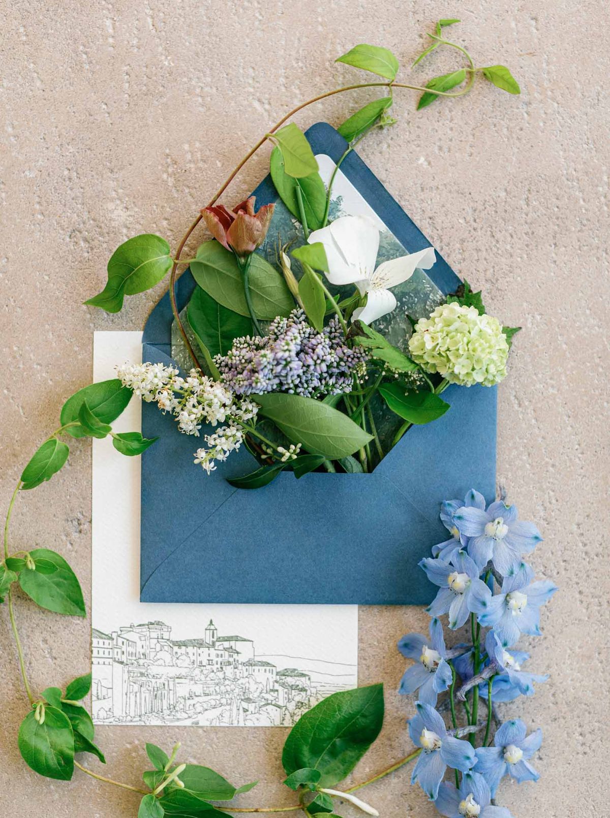 Wedding stationery flat lay with teal envelope, white cosmos, lavender blooms, and green foliage on stone