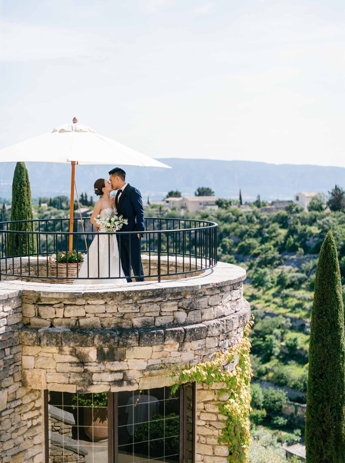 Couple kissing on circular stone terrace with white umbrella, cypress trees, and Tuscan valley panoramic view below