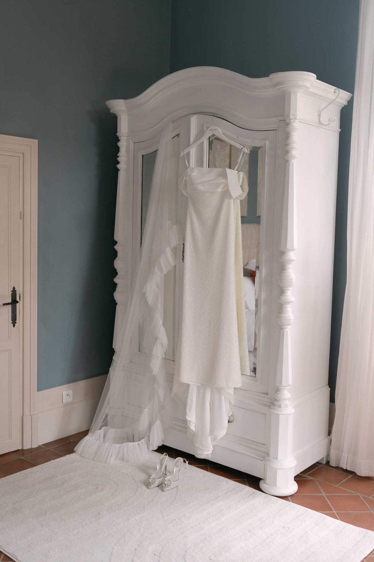 A getting-ready detail shot taken indoors showing a bride's ivory slip-style wedding dress with spaghetti straps hanging from a white ornate French armoire with mirrored panels and carved detailing. A long, ruffled-edge cathedral veil in ivory-white is draped over the left side of the armoire, pooling onto the floor. A pair of silver strappy heeled sandals with embellished detailing sits on a white textured rug at the base of the armoire. The room features teal-blue painted walls, terracotta tile flooring, and a white panelled door with black iron hardware, suggesting a classic French château interior. The composition is a medium-wide shot capturing the full length of the dress and veil against the room setting.