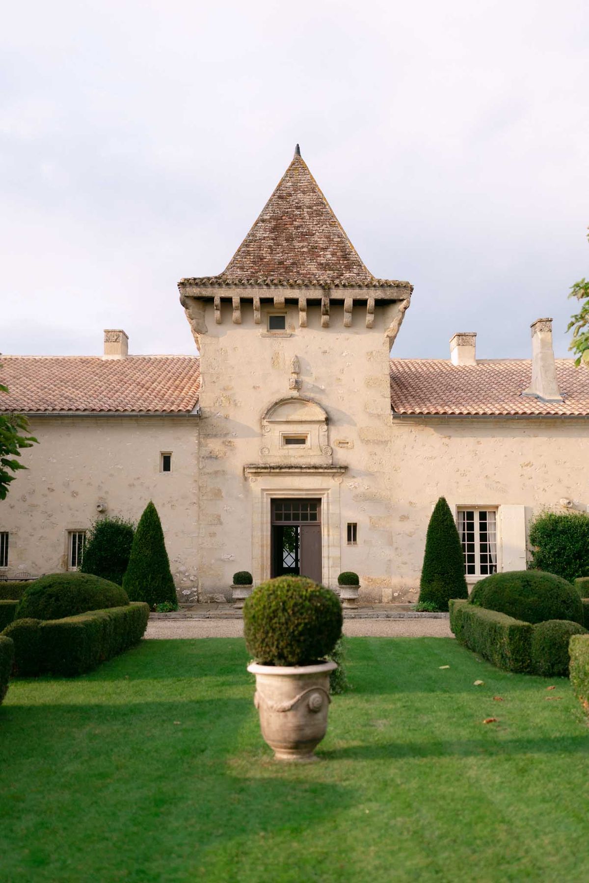 Chateau with central tower, arched entrance, conical topiary, and stone urn planter on formal lawn