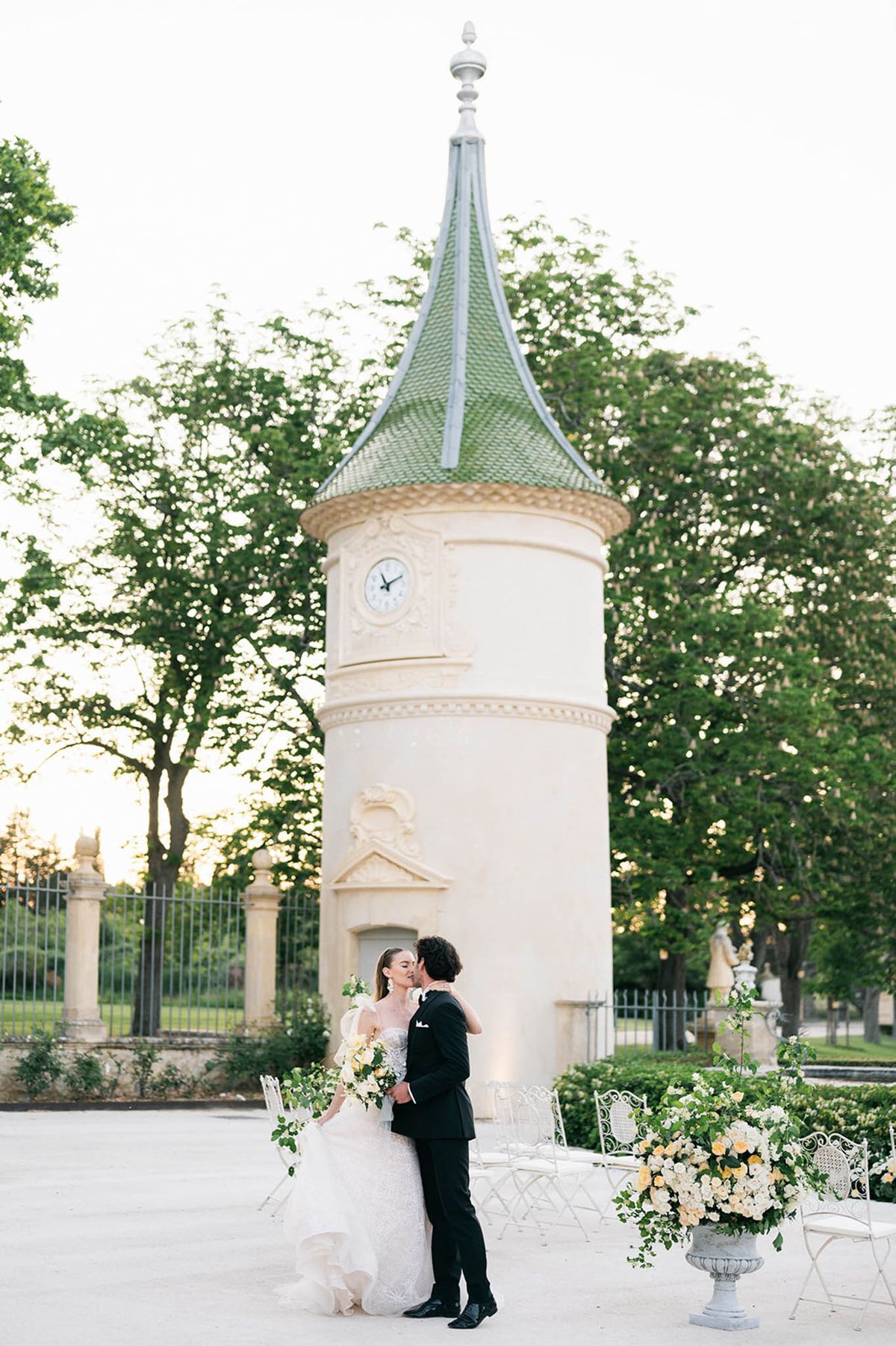Bride and groom kissing before chateau round tower with green roof, white rose urns, and bistro chairs