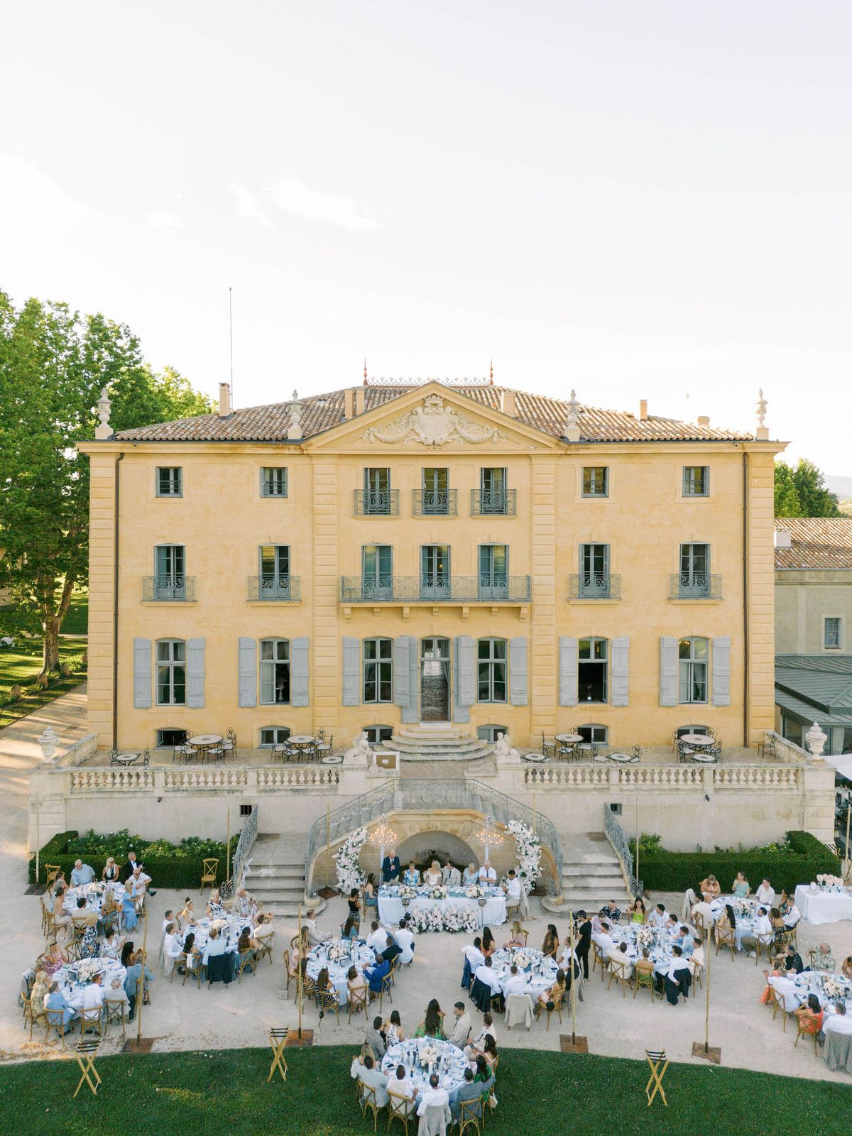 An aerial wide shot of an outdoor wedding reception taking place on the grounds of a classic French château. The three-story manor features warm yellow ochre stone facade, pale grey-blue shutters, ornate pediment detailing, wrought iron balconies, and a terracotta tile roof. Approximately 80–100 guests are seated at round tables with white linens, arranged across a gravel courtyard directly in front of the château's grand stone staircase and balustraded terrace. The sweetheart table is positioned centrally at the base of the staircase, framed by a large white floral arch and flanked by candelabras, with the couple and a small group of guests visible. Table centerpieces appear to feature white floral arrangements, and gold crossback chairs are used throughout. Small gold floor-standing candelabras are spaced between tables. The decor palette is white, gold, and soft blue, consistent with a classic, formal aesthetic. The image is taken during golden hour, with warm evening light bathing the facade. Potential venue feature image.
