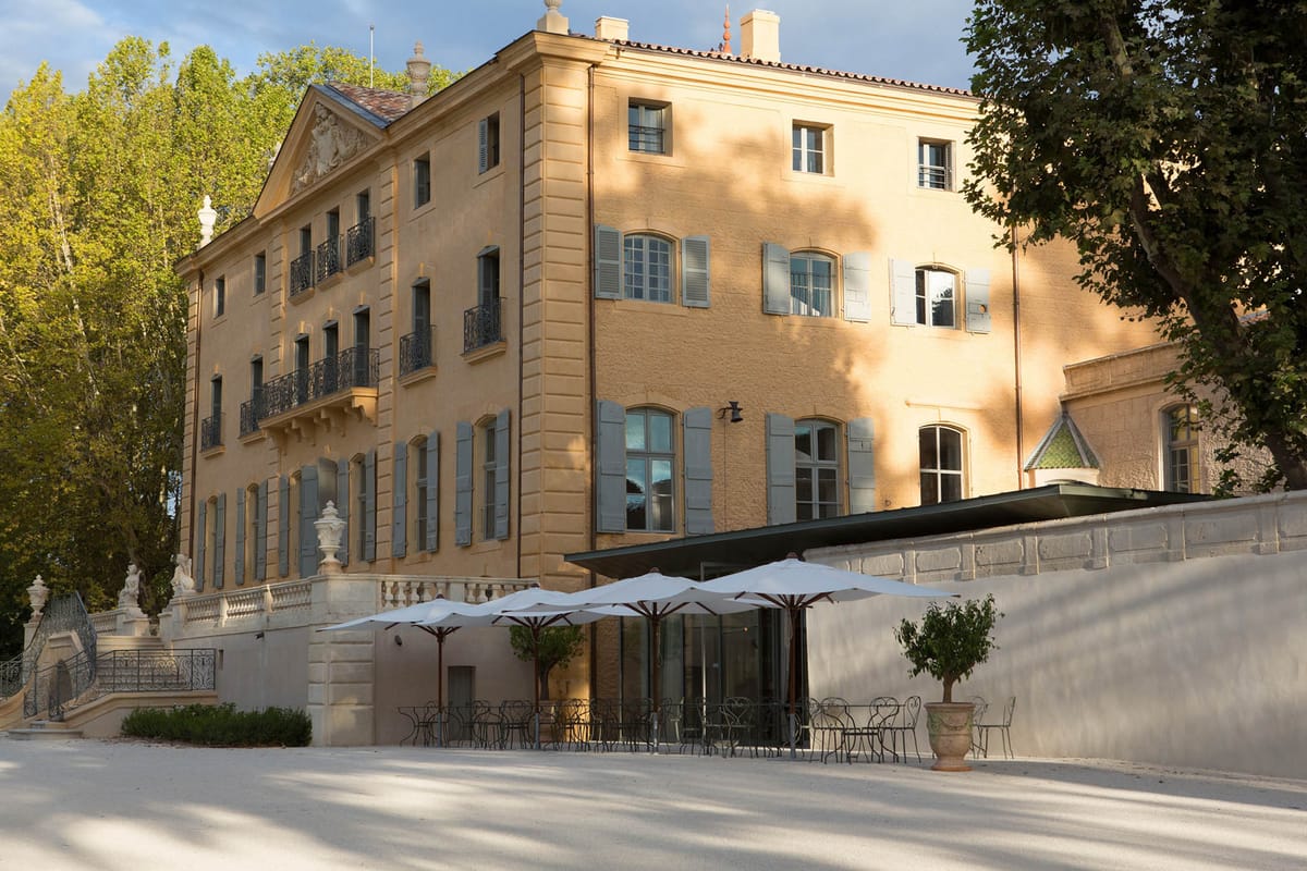 An exterior wide shot of a large French château-style manor house photographed in warm late-afternoon light. The three-storey building features ochre-yellow rendered walls, pale blue-grey shutters, wrought-iron juliet balconies, and classical stone detailing including a decorative pediment, balustrade, and sculptural urns on a grand staircase. At ground level, a covered terrace with a dark canopy extends from the building, set with dark metal bistro chairs and tables beneath a row of large white market umbrellas on dark wooden poles. Two clipped topiary trees in terracotta pots flank the terrace area on a pale gravel forecourt. No people are visible in the image. Potential venue feature image.