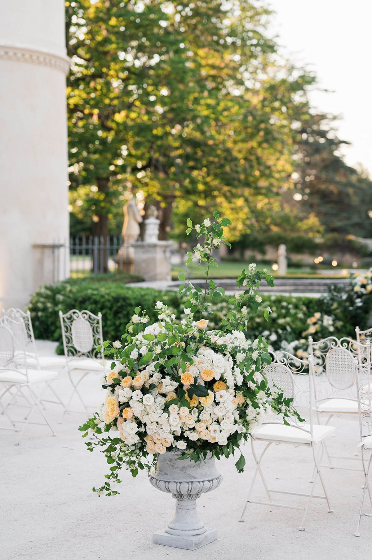 Stone urn with apricot and cream garden roses beside wrought-iron chairs on parterre terrace at estate