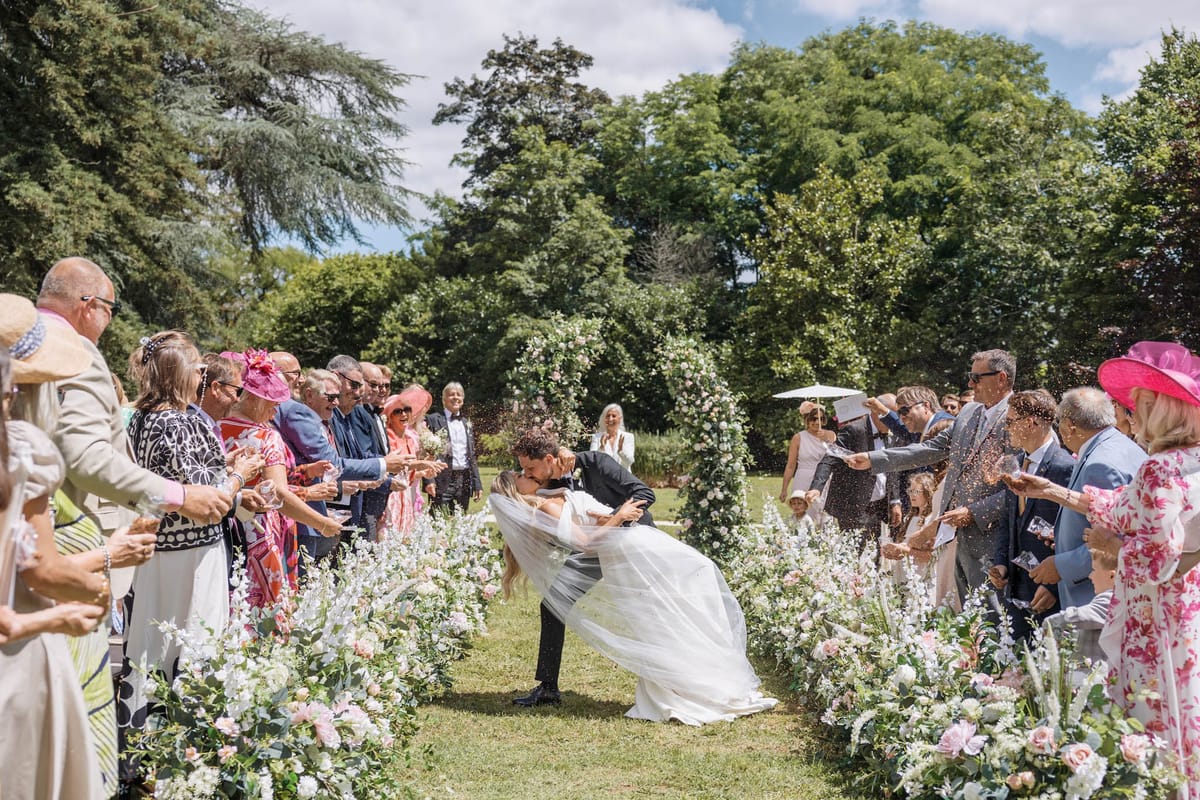 The couple shares a dip kiss while walking back down the aisle following an outdoor garden ceremony, surrounded by approximately 40 guests on either side who are throwing confetti. The bride wears a white gown with a long flowing veil, and the groom is in a dark suit. The aisle is lined with low ground-level floral arrangements featuring blush pink roses, white delphinium, and trailing greenery, with a large floral arch of blush and white roses visible at the far end of the aisle. Guests are dressed in a mix of formal attire including brightly colored fascinators and hats in pink and coral tones, navy and blue suits, and floral dresses. The setting is a manicured garden lawn with mature trees in the background, photographed in bright midday sunlight. The shot is taken from a wide mid-level angle looking straight down the aisle, capturing both rows of guests and the full length of the floral aisle décor.