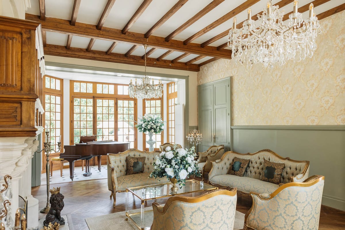 An interior reception or lounge area of what appears to be a French château, decorated for a wedding event. The room features gold-and-cream damask wallpaper, sage green wainscoting and paneled doors, exposed dark wood ceiling beams, and two crystal chandelier pendant lights. The seating area consists of Louis XVI-style sofas and armchairs upholstered in gold brocade fabric with gilt frames, arranged around a gold and glass coffee table topped with a large floral centerpiece of white roses and green foliage in a white urn-style vase. A second tall white pedestal vase with a full arrangement of white roses and greenery stands near the entrance to an adjoining sunroom, where a grand piano is visible against a backdrop of floor-to-ceiling French windows. A white marble fireplace with candelabras and a bronze lion figurine is visible on the left edge of the frame. No people are present. Wide interior shot. Potential venue feature image.