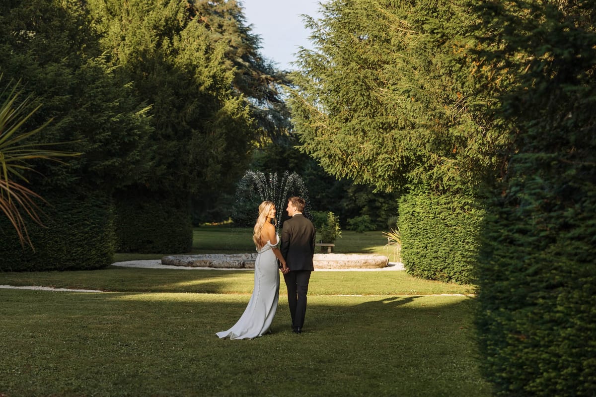 A couple portrait taken outdoors in a formal garden setting, photographed as a medium-wide shot from behind. The bride wears a fitted ivory off-the-shoulder gown with a low open back, button detail down the spine, and a short train; she turns her head back toward the camera while holding the groom's hand. The groom is dressed in a dark charcoal suit and faces away from the camera. The two are standing on a manicured lawn flanked by tall trimmed hedges and mature conifer trees, with a stone fountain basin and water jets visible directly behind them. The overall styling is clean and modern, with warm golden-hour light casting long shadows across the lawn.