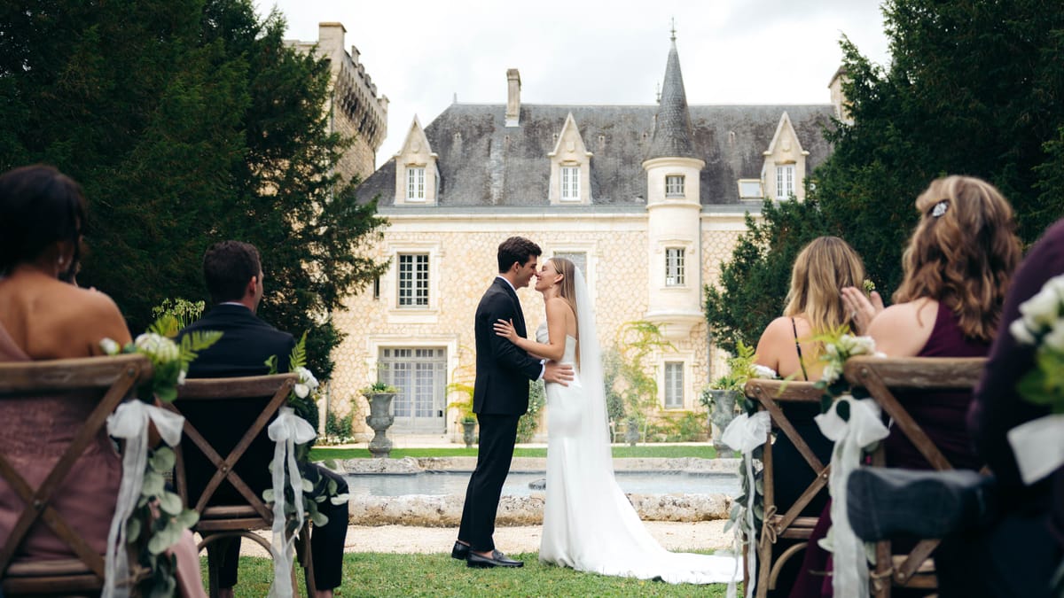 An outdoor wedding ceremony first kiss moment captured in a wide shot, with the couple standing at the end of the aisle in front of a classic French château built in pale limestone with conical turrets and a slate roof. The groom wears a dark navy suit and the bride wears a sleek, minimalist white slip-style gown with a long white veil. A rectangular stone fountain pool is visible between the couple and the château façade. Guests are seated in rustic cross-back wooden chairs arranged on either side of the grass aisle, adorned with white ribbon and small white floral and greenery clusters. Guests visible in the foreground include women in mauve and deep burgundy/plum dresses. The overall decor palette is white and green against the natural stone backdrop, with a classic, understated styling approach. Potential venue feature image.